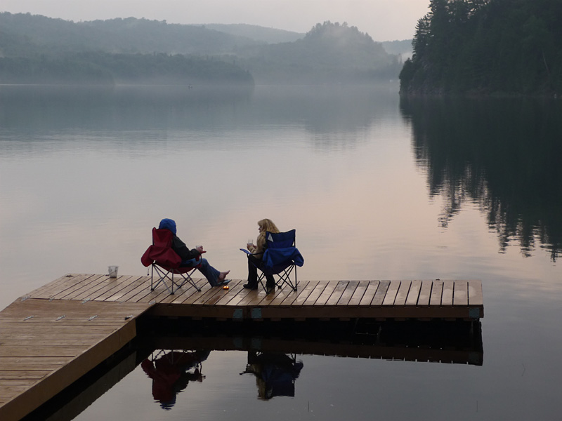This is a photo of two people on the dock drinking coffee with mist coming off the late and you can just see the water and shore on the other side